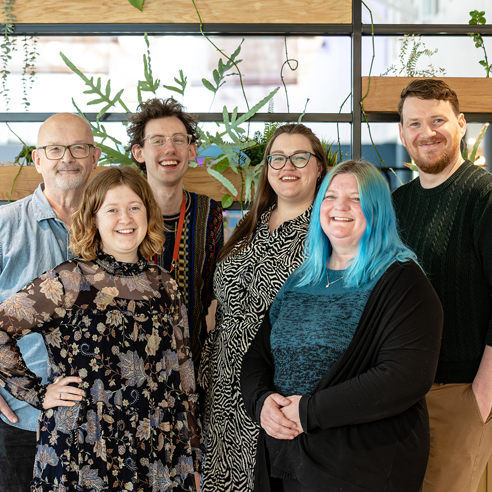 The All In team smile as they pose for a photo in a bright environment with plants and modern decor.