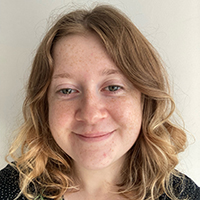 Gemma, a white woman with auburn shoulder-length wavy hair and freckles smiles at the camera. She wears a black and white spotted top.