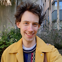 Jamie, a white person with brown hair, stands in a garden and smiles at the camera. They wear a bright yellow coat over a multi-coloured jumper.