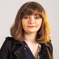 Joanne, a white woman with shoulder length brown hair and a fringe poses for the camera. She wears a black leather jacket and a floral dress.