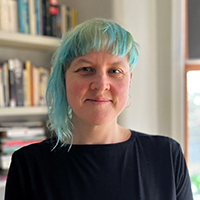 Kelly, a white woman with blue hair that is long on one side. She wears a black t shirt and stands in front of bookshelves.