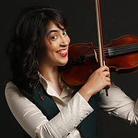 Lilit, an amputee in her 30s, holds her violin up about to play, while smiling at the camera. She has dark brown shoulder-length wavy hair and wears a dark green waistcoat over a white shirt with decorative cuffs.