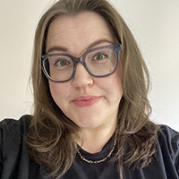Nicola, a white woman with shoulder length brown hair and purple glasses smiles at the camera. She wears a black t-shirt with ‘Rocket Man’ in white text.