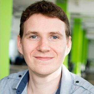 Phil, a white man with short brown hair smiles at the camera. He wears a blue, open neck shirt and stands in an office with green pillars.