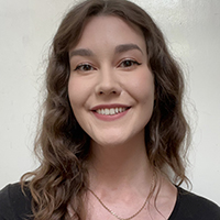 Ruby, a white woman with long brown wavy hair and winged eyeliner smiles at the camera. She wears a black long-sleeved top and a gold necklace