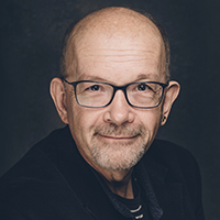 Tim, a white, bald man with a grey moustache and goatee poses for the camera. He wears a navy jacket over a navy and white striped t-shirt.