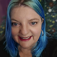 Roberta, a white woman with dyed blue hair, smiles at the camera. She wears big silver hooped earrings and a teal wool jumper.