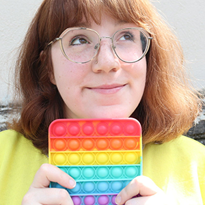 Al is a white woman with freckles, shoulder length auburn hair and wearing metal framed glasses. She holds up a rainbow-striped bubble fidgeter and smiles off-camera.