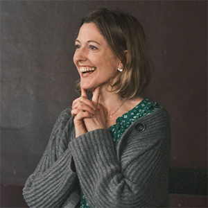 Jody is a white woman with shoulder-length dark brown hair streaked with blonde. She smiles off camera, with her hands clasped under her chin. She is wearing a green floral dress and grey knitted cardigan.