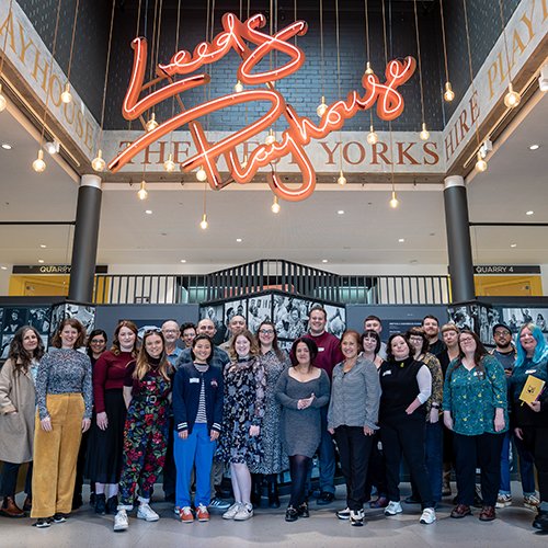 A big, smiling group photo of the All In team with members of staff from the pilot organisations and suppliers working on the project. They gather in the lobby of Leeds Playhouse with a big red neon sign hanging overhead.